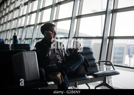 Portrait of young handsome man wearing casual style clothes sitting on the bench in modern airport using smartphone. Passenger travelling with luggage bag making call, while waiting for his flight Stock Photo
