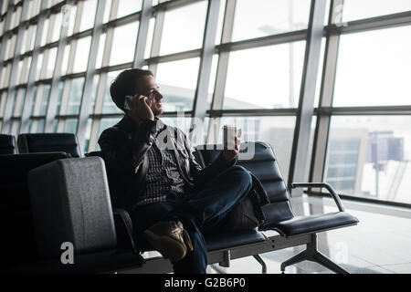 Portrait of young handsome man wearing casual style clothes sitting on the bench in modern airport using smartphone. Passenger travelling with luggage bag making call, while waiting for his flight Stock Photo