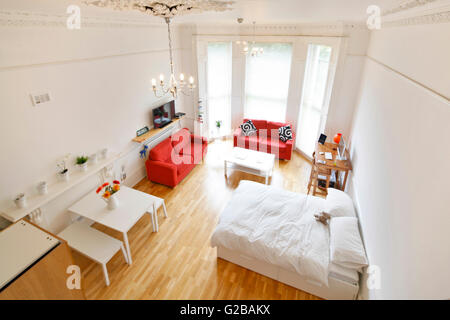 Pembridge Square, Notting Hill. View of a spacious, contemporary studio apartment with a bed with white linen and orange couches. High ceiling and large windows. Stock Photo