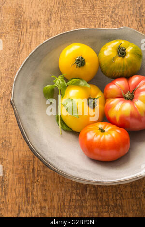 Multicolored heirloom tomatoes and basil in a ceramic bowl on a rustic wooden surface. Stock Photo