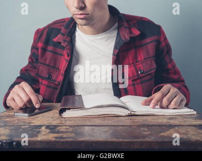 A young man is sitting at a wooden desk with a smartphone and a notebook Stock Photo
