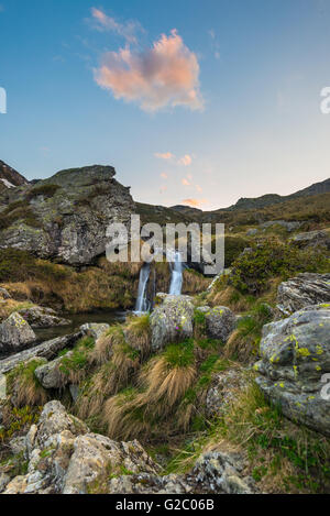 Blurred flowing waterfall in the Alps with colorful sky and clouds at twilight, among rocks, boulders and grass. Little stream i Stock Photo