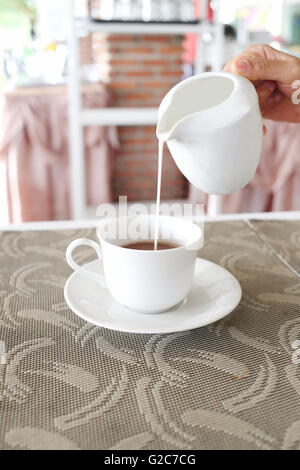 Women's hands are pouring milk from a white jug into a coffee cup on table food. Stock Photo