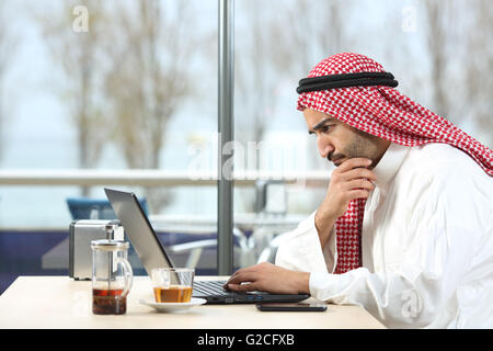 Side view of an arab saudi man worried working with a laptop in a coffee shop interior with the terrace in the background Stock Photo