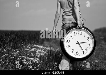 Horizontal rear view of a young woman carrying a big clock on a field Stock Photo