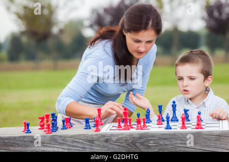 Elegant young boy in white shirt and his mother learning to play chess with blue and red chess pieces on wood table in the park Stock Photo