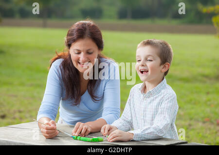 Young woman teacher teaches little young boy in white shirt painting with the brush to make artisanal pasta beads Stock Photo