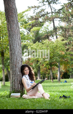 Young woman asian with long curly hair laying on grass against a tree writing in the park Stock Photo