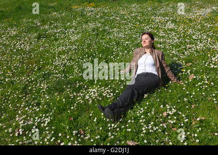 Relaxing young woman in pants and leather jacket enjoying spring, lying on grass and spring flowers Stock Photo