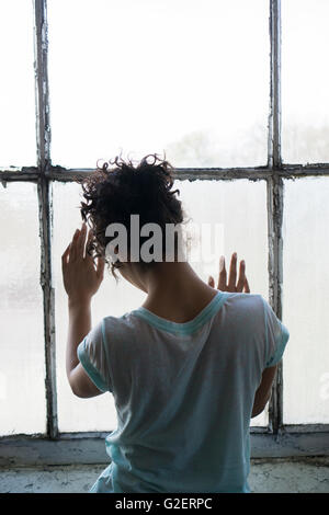 Rear view of a young woman standing by the window hands touching the glass Stock Photo