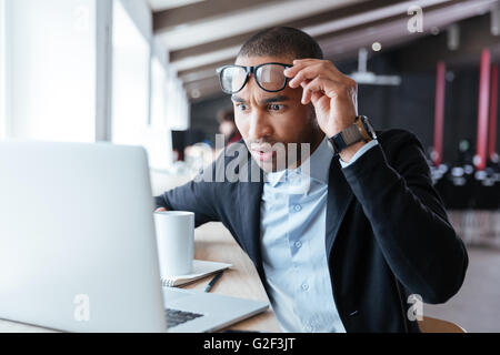 Businessman receiving negative news, touching his glasses, being mad, upset and surprised in front of laptop Stock Photo