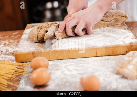 Closeup of hands of man baker cutting bread on wooden board on the kitchen Stock Photo