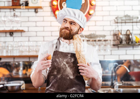 Pensive bearded baker holding eggs and bread and thinking on the kitchen