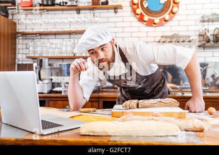 Thoughtful bearded baker cutting bread and using laptop on the kitchen Stock Photo