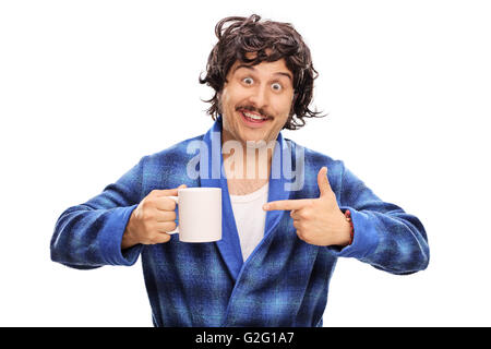 Excited young man in a blue bathrobe holding a ceramic cup and pointing to it with his finger isolated on white background Stock Photo