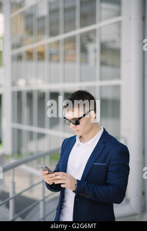 Portrait of a young businessman seeing on his smartphone on the street. Man with dark sunglasses. Stock Photo