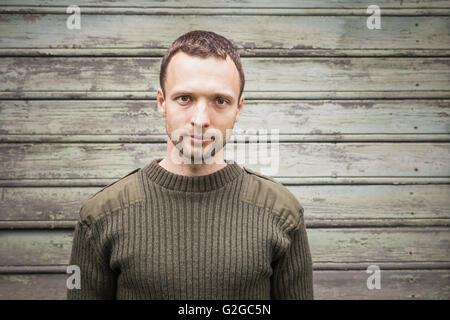 Young handsome Caucasian man in military green sweater. Outdoor close up portrait over green rural wooden wall Stock Photo