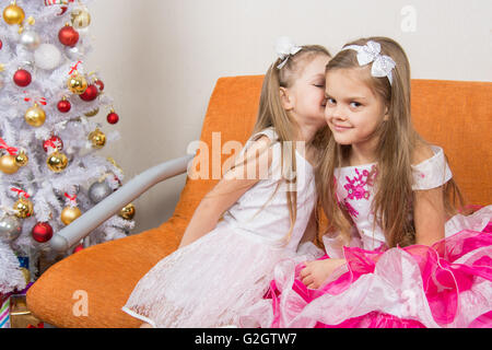 Two girls in beautiful dresses whispering sitting on couch at Christmas tree Stock Photo