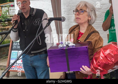 Harmondsworth, London, UK. 30th May, 2016 A woman brings a large purple  wrapped present for Heathrow Airpots to the microphone at the 'celebration' of Heathrow's 70th birthday by HACAN and  anti-3rd runway campaigners in Harmondsworth. She says the box contains some of Heathrow's many broken promises to the people of the area over the years.. Credit:  Peter Marshall/Alamy Live News Stock Photo