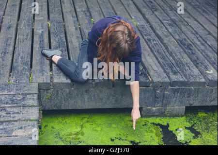 A young woman is sitting on a wooden deck by a pond in a forest and is pointing at something in the water Stock Photo
