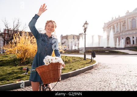 Cheerful lovely young woman with bike standing and waving her hand on the street Stock Photo