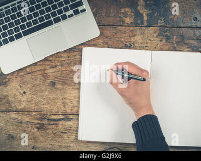 Overhead shot of a male hand writing in a notepad. There is a laptop on the table as well. Stock Photo