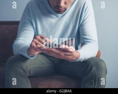 A young man is sitting on an old antique sofa and is using a smart phone Stock Photo