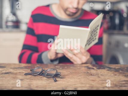 A young man is reading a book in a kitchen. Focus on his reading glasses on the table in front of him. Stock Photo