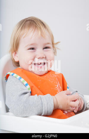 blonde caucasian baby seventeen month age orange bib grey sweater on white high chair laughing looking at camera eating Stock Photo