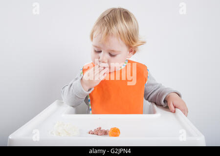 blonde caucasian baby seventeen month age orange bib grey sweater eating meal with her hand in white high-chair Stock Photo