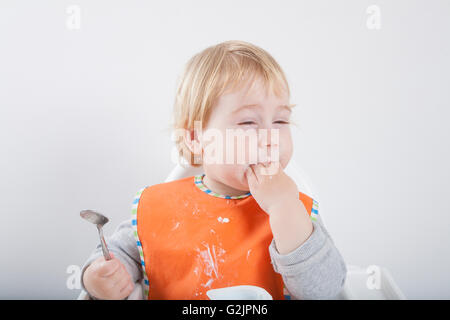 blonde caucasian baby seventeen month age orange bib grey sweater eating meal with her hand in white high chair Stock Photo