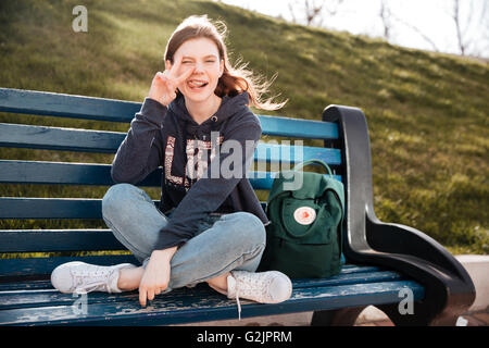 Funny playful young woman winking and showing peace sign sitting on the bench Stock Photo