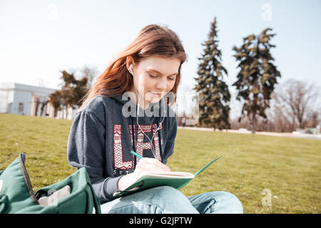 Smiling cute young woman in earphones listening to music and writing in notebook sitting on the lawn Stock Photo