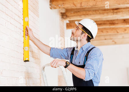 Close-up portrait of a builder holding construction level against the wall and standing on the ladder indoors Stock Photo