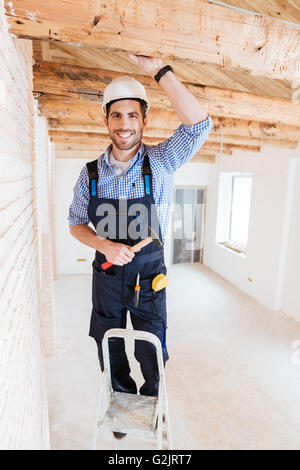 Smilling worker standing on the ladder and holding a hammer indoors Stock Photo