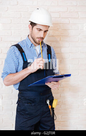 Pensive young construction worker thinking about something using clipboard while working indoors Stock Photo