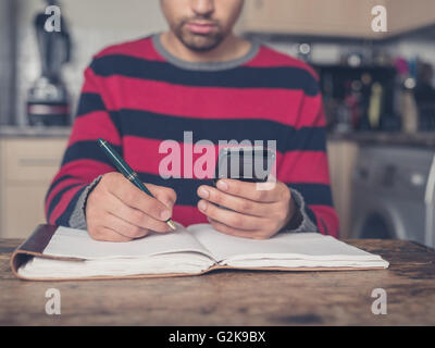 A young man is sitting at a table in a kitchen with a notebook and is using a smart phone Stock Photo