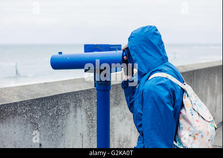 A person wearing a blue jacket and hood is using a telescope on the coast Stock Photo