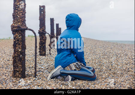 A young woman is sitting on the shingle by a rusty fence on the beach Stock Photo