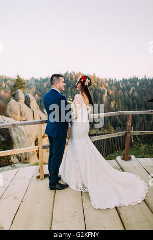 Elegant groom in stylish blue suit lovingly holding his charming white dressed bride standing on wooden platform with majestic mountain rocky landscape as background. Back view Stock Photo