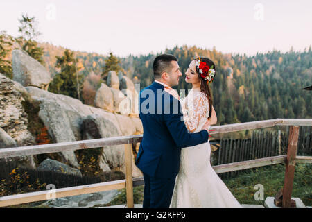 Elegant groom in stylish blue suit lovingly holding his charming white dressed bride standing on wooden platform with majestic mountain rocky landscape as background Stock Photo