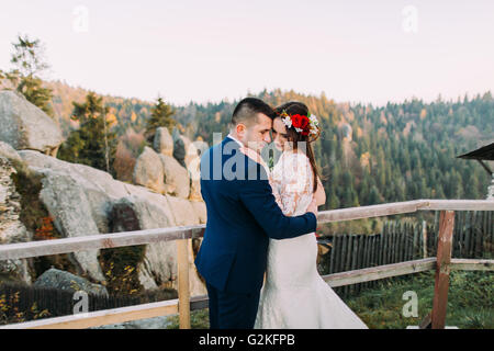 Elegant groom in stylish blue suit lovingly holding his charming white dressed bride standing on wooden platform with amazing mountain rocky landscape as background. Back view Stock Photo