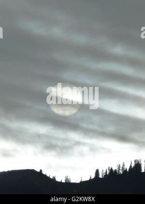 Setting sun behind a veil of clouds with a wooded mountain ridge silhouetted against the light Stock Photo