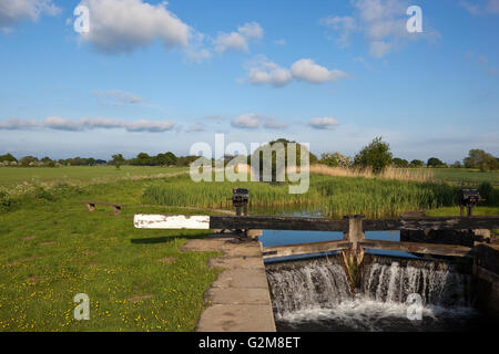 Water pouring through canal lock gates beside a rural towpath with benches and picnic area in an agricultural landscape in May. Stock Photo