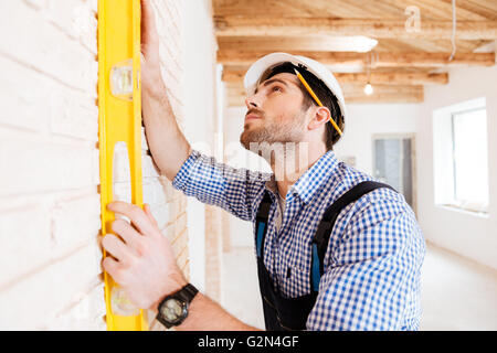 Concentraated builder in uniform holding a level against the wall indoors Stock Photo