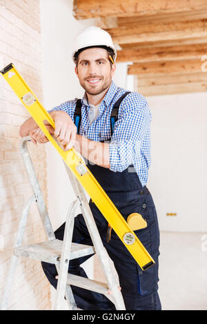 Close-up portrait of a smiling worker standing on the ladder and holding construction yellow level inoors Stock Photo