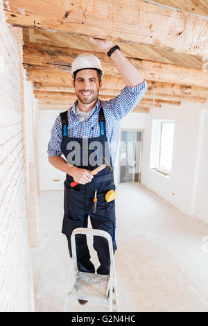 Smilling worker standing on the ladder and holding a hammer indoors Stock Photo