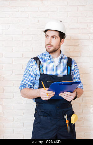 Young handsome builder thinking about something and holding a clipboard in his hands while working indoors Stock Photo