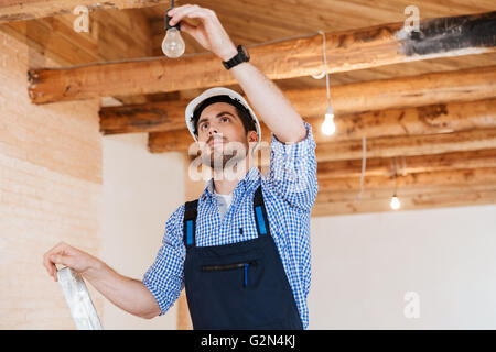 Handsome young builder fixing socket using ladder indoors Stock Photo