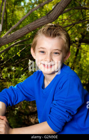 A young boy sits in a wooded area and smiles for a portrait.  He has blond hair and blue eyes and is wearing a bright blue shirt Stock Photo